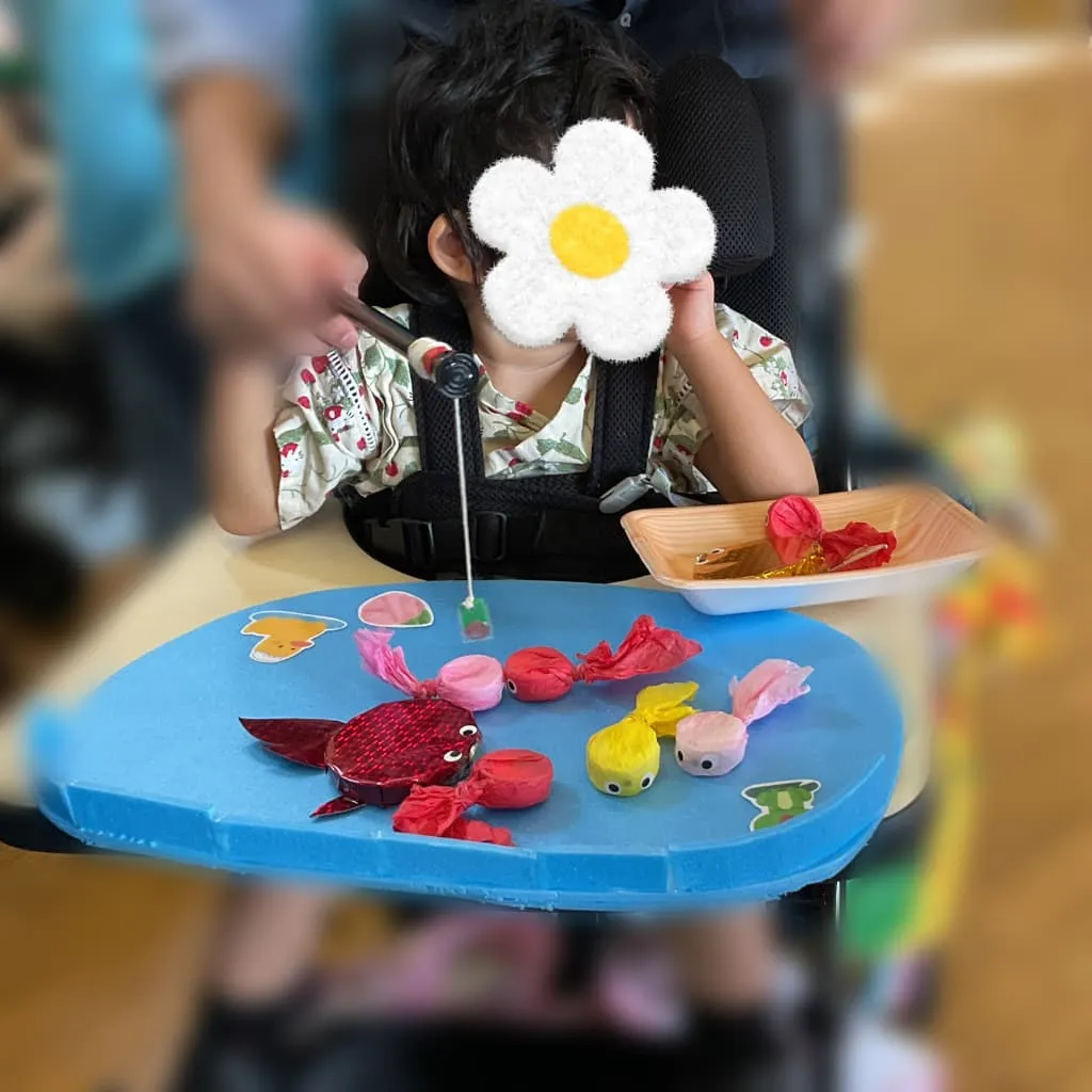 室内で車椅子に座り、大人と一緒におもちゃの釣り竿をもちながら、色とりどりの魚を釣ろうとする女の子