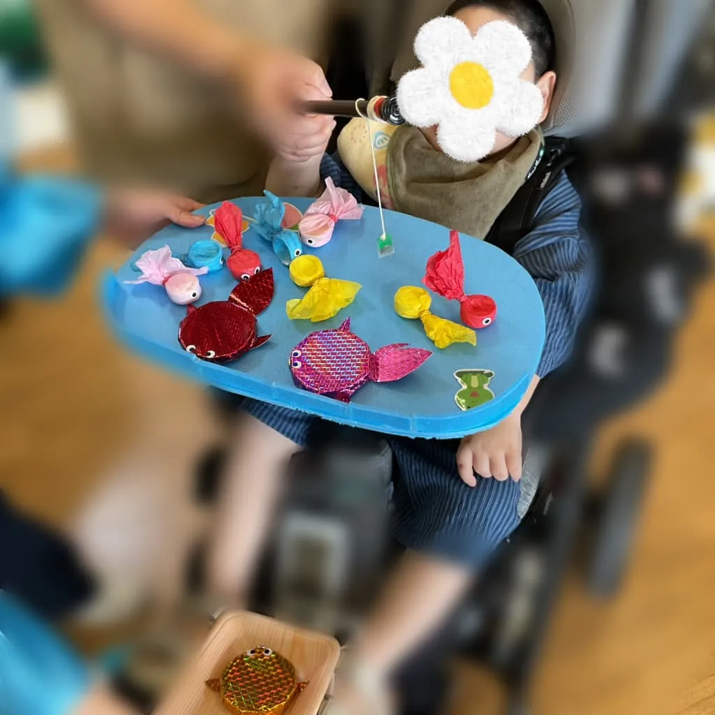 室内で車椅子に座り、大人と一緒におもちゃの釣り竿をもちながら、色とりどりの魚を釣ろうとする男の子