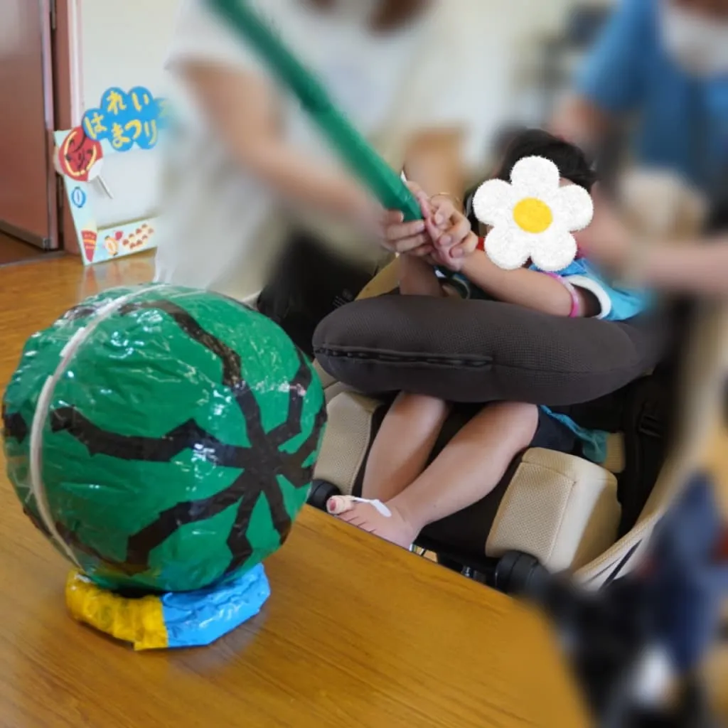 室内で車椅子に座り、介助を受けながら緑色の棒でスイカ割りを楽しむ子ども